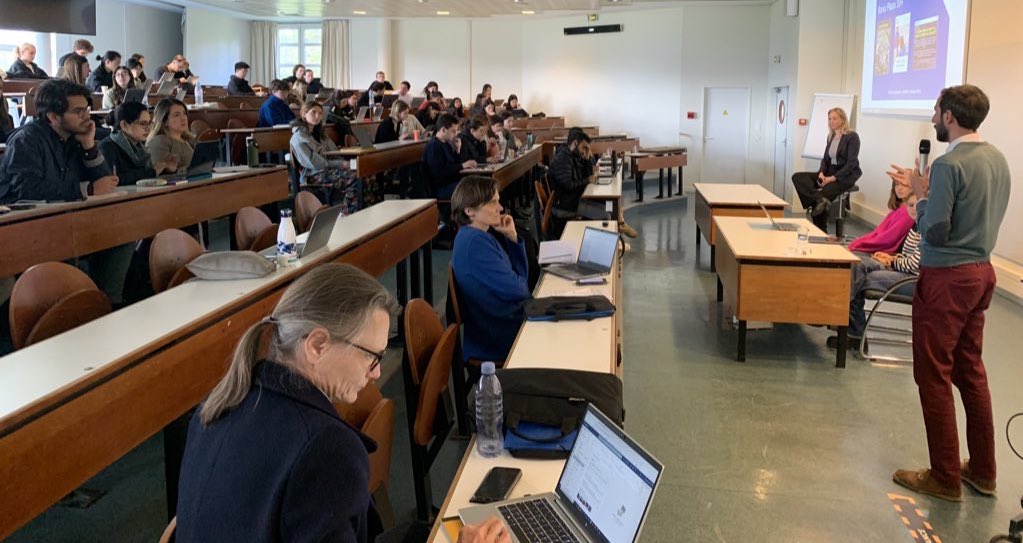 A classroom filled with students sitting at desks arranged in rows, listening to a person speaking at the front of the room. The students are using laptops and taking notes