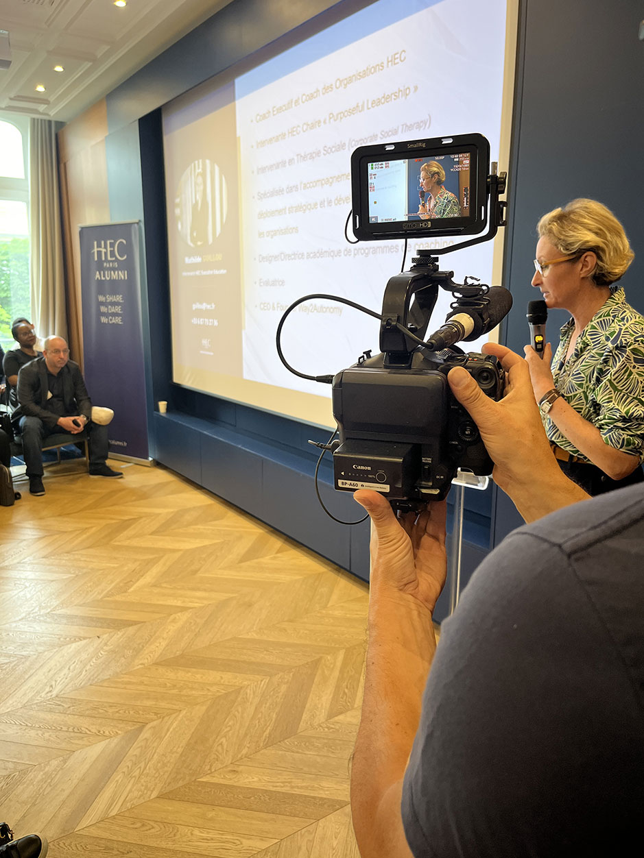 Une femme présente devant un écran lors d'un événement HEC Alumni. Un caméraman filme la scène, tandis que plusieurs personnes sont assises et écoutent attentivement en arrière-plan.