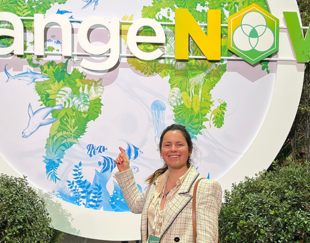 A smiling woman pointing at a sign with the text 'ChangeNOW' and illustrations of nature and marine animals in the background, surrounded by greenery