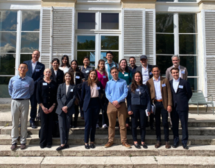 A group photo of around 30 people attending a workshop at HEC, posing on the steps of a large, elegant building with white walls and multiple windows. The attendees are casually dressed and arranged in a relaxed manner, some sitting and others standing, with a sunny blue sky and green trees in the background.