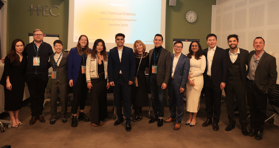 A group of twelve people posing together in front of a screen displaying 'HEC Paris Le Château'. They are all dressed in business or smart casual attire, smiling and standing in a row. The group includes both men and women of diverse backgrounds