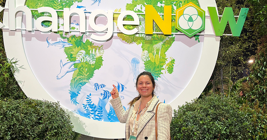 A smiling woman pointing at a sign with the text 'ChangeNOW' and illustrations of nature and marine animals in the background, surrounded by greenery