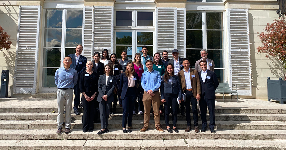 A group photo of around 30 people attending a workshop at HEC, posing on the steps of a large, elegant building with white walls and multiple windows. The attendees are casually dressed and arranged in a relaxed manner, some sitting and others standing, with a sunny blue sky and green trees in the background.